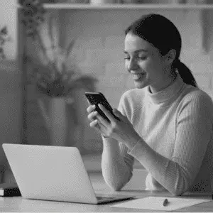 Contáctanos muestra la foto de una joven mujer sentada ante una laptop abierta y sonriente manipulando en su móvil.